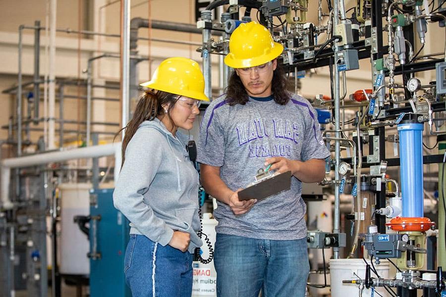 Two 火博体育 students wearing hard hats checking a checklist in front of a series of pipes